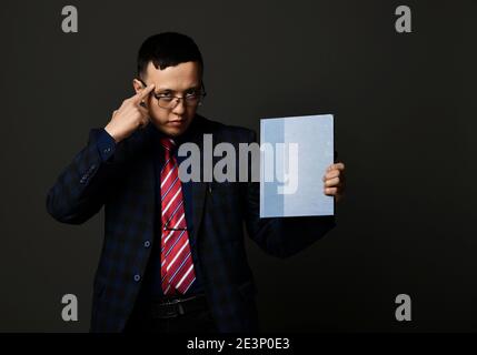 Giovane uomo d'affari asiatico serio in piedi tenendo carta blu vuota dentro mano e puntando alla testa che spinge a pensare e ad avere idee Foto Stock