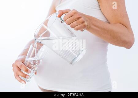 Donna incinta che versa acqua dal vaso al bicchiere su sfondo bianco Foto Stock