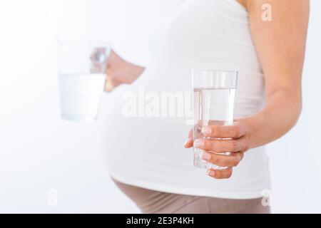 Donna incinta che tiene un bicchiere pieno d'acqua e il vaso su sfondo bianco Foto Stock