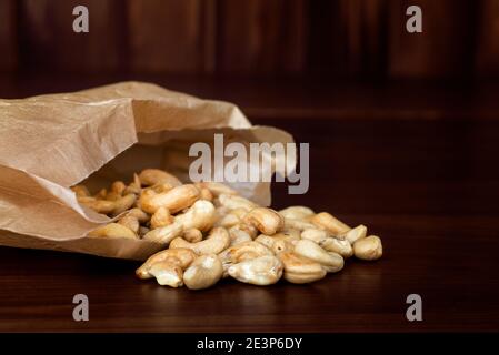 Anacardi sparsi in un sacchetto di carta su sfondo di legno rustico scuro. Noci di anacardi, snack Foto Stock