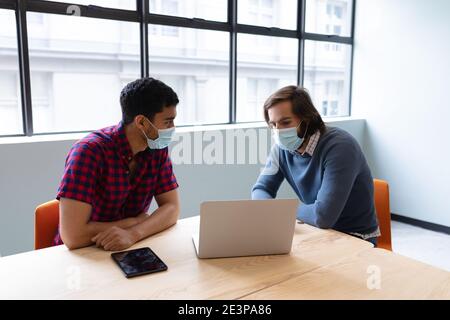 Due uomini d'affari diversi che indossano maschere di fronte utilizzando il computer portatile e discutere in ufficio creativo Foto Stock