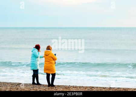 Due donne in abiti luminosi sono in piedi sulla spiaggia, ammirando la vista. Vista posteriore. Il concetto di attività ricreative all'aperto. Foto Stock