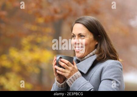 Donna felice che beve caffè guardando via in inverno fuori dentro un parco Foto Stock