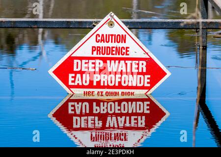 Segnale di avvertimento di acqua profonda. Parte del segno viene sommersa. Il cartello è in inglese e francese. Il riflesso del segno è nell'acqua sottostante. Foto Stock