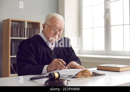 Giudice serio saggio maturo in abito seduto alla scrivania dentro courtroom e la lettura di un libro Foto Stock