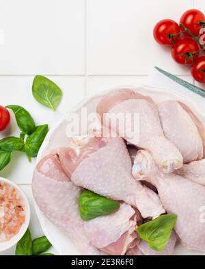 Carne di pollo cruda, pronta per grigliare o grigliare le gambe, con pomodori, erbe e spezie su sfondo bianco chiaro del tavolo da cucina. Vista dall'alto. Foto Stock