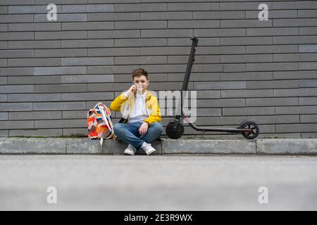 Adolescente moderno con zaino e scooter elettrico seduto sul marciapiede e parlare al telefono Foto Stock