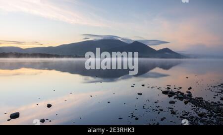 Il Massiccio di Skiddaw si riflette nelle acque calme del Lago di Bassenthwaite durante un'alba a marcia pastello con accenni di nebbia che galleggia sull'acqua. Foto Stock