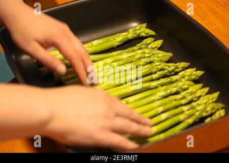 Le mani delle donne mettono un fascio di asparagi biologici freschi su un vassoio da forno nero Foto Stock