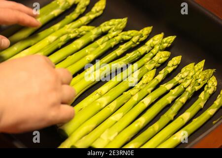 Le mani delle donne mettono un fascio di asparagi biologici freschi su un vassoio da forno nero Foto Stock