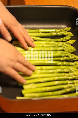 Le mani delle donne mettono un fascio di asparagi freschi su una griglia da forno nera e cucinano a casa Foto Stock