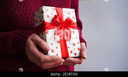 Il concetto di banner di San Valentino, una donna in un pullover borgogna contiene una confezione regalo avvolta in carta bianca con cuori rossi e legata con un arco di nastro rosso, Foto Stock