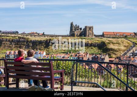 Un uomo e una donna seduti su una panchina guardando verso il porto e oriente scogliere di Whitby,North Yorkshire compreso l'abbazia Foto Stock