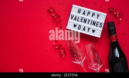 Concept banner giorno di San Valentino.Board con testo Happy valentines day,serpentina,champagne,bicchieri e confetti a forma di cuore su sfondo rosso, vista dall'alto, Foto Stock