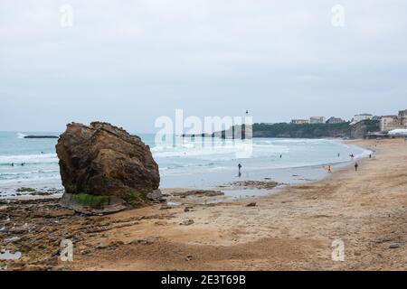 Biarritz, Francia. Vista di Big Beach con surfisti (irriconoscibili) in acqua, rocce, faro e edifici della città nel nuvoloso giorno di primavera. Foto Stock