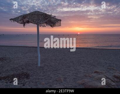 Alba colorata sulla spiaggia vuota di Santa Maria Navarrese con ombrellone in legno, sole rosso che si alza dal mare e nuvole rosa blu e arancio Foto Stock