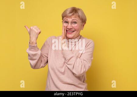 Donna caucasica matura che punta con indice dito da parte sussurrando segreto dietro la sua mano isolato su sfondo giallo parete. Foto Stock