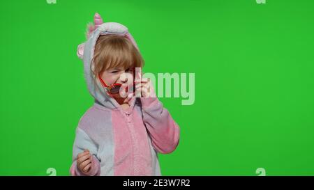 Piccolo biondo bambino danzare, celebrare, fare i volti, inarcare in costume unicorno con occhiali da sole su sfondo verde chiave Chroma. Ritratto di animatore ragazzino in pigiami unicorno. Spazio di copia Foto Stock