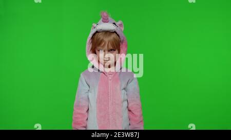 Giovane piccolo biondo sconvolto bambino di 5-6 anni facendo volti in costume unicorno su sfondo verde chiave cromatica. Ritratto di animatore ragazzino in pigiami unicorno. Spazio di copia Foto Stock