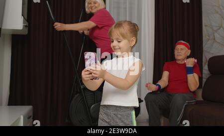 Anziano anziano anziano coppia uomo donna utilizzando orbitrek facendo sollevamento pesi sport manubri esercizi di formazione a casa. Bambina che fa selfie videochiamata sul telefono cellulare per mostrare come cool nonni sono Foto Stock