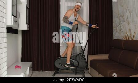 Divertente uomo atleta sportivo ragazzo anni '80 con barba rende allenamento fitness cardio su orbitrek a casa. Giovane uomo hipster di stile che pratica lo sport, divertendosi a girare intorno. Concetto di parodia fitness Foto Stock