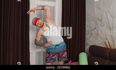 Il simpatico atleta sportivo degli anni '80 con barba fa stretching in allenamento a casa. Ritratto di un giovane hipster di stile che pratica lo sport, divertendosi, si diverte. Concetto di parodia fitness Foto Stock