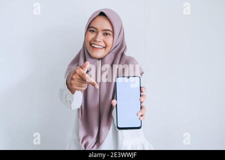 Giovane islam asiatico donna che indossa il copricapo sta puntando il dito sullo schermo bianco del telefono con sorriso e sensazione felice. Donna indonesiana sullo sfondo grigio Foto Stock