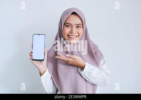 Giovane islam asiatico donna che indossa il copricapo sta puntando il dito sullo schermo bianco del telefono con sorriso e sensazione felice. Donna indonesiana sullo sfondo grigio Foto Stock