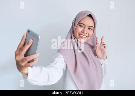 Giovane islam asiatico donna con foulard è sorridente e selfie scattare foto con il suo telefono cellulare. Donna indonesiana su sfondo grigio. Foto Stock