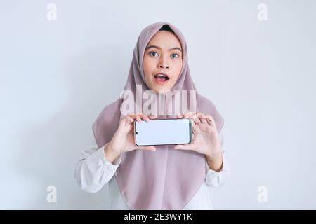 Giovane islam asiatico donna che indossa il copricapo è mostrato sullo schermo bianco del telefono cellulare in vista orizzontale con shock e sensazione felice. Donna indonesiana o Foto Stock