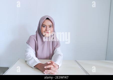 Giovane islam asiatico donna che indossa il copricapo è seduta sul tavolo con le mani e il sorriso. Islam donna indonesiana su sfondo grigio. Foto Stock