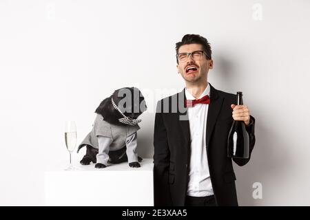 Uomo triste piangendo e bevendo champagne dalla bottiglia, mentre pug in simpatico costume da festa fissando confuso, in piedi su sfondo bianco Foto Stock