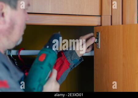 Servizi di assemblaggio e riparazione di mobili. Un uomo in grigio e rosso abiti speciali per lavorare con un cacciavite viti su una cerniera per porta di mobili. Primo piano, s Foto Stock