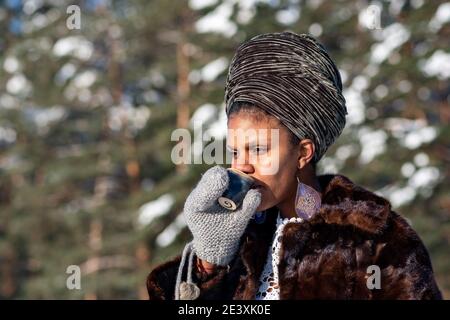 Vilnius, Lituania - Gennaio 20 2021: Bella ragazza con cappotto di pelliccia bere caffè caldo o tè sulla neve su un lago ghiacciato in inverno con alberi di foresta Foto Stock