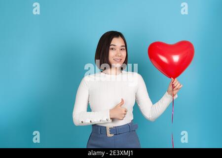 Sorridente, felice giovane donna asiatica tiene un pallone volante a forma di cuore e dà un pollice su isolato su uno sfondo blu Foto Stock