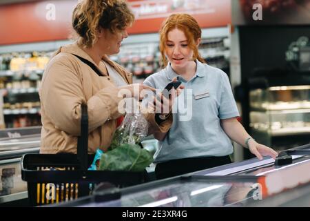 Operaio del negozio di alimentari che assiste il cliente femminile. Cliente femminile che mostra il suo telefono cellulare all'assistente del negozio e chiede il prodotto al supermercato. Foto Stock