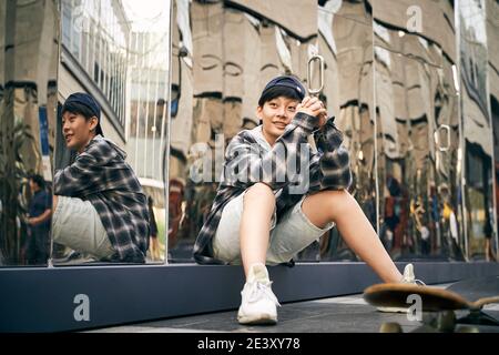 teenage asiatico bambino skateboarder prendere una pausa seduta a terra sorridente Foto Stock