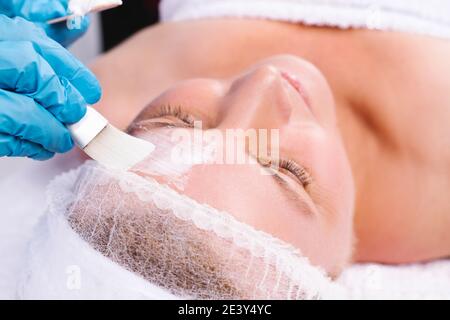 Una foto ingrandita di una donna caucasica anziana con un fasciatesta e le mani guintate di estetista applicano la maschera facciale con un pennello cosmetico. Foto Stock