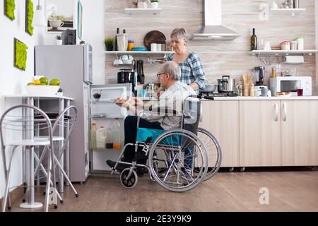 Uomo anziano in sedia a rotelle che prende il cartone delle uova dal frigorifero che aiuta la moglie in cucina. Donna anziana che prepara la colazione per il marito handicappato, che vive con l'uomo con disabilità motorie Foto Stock