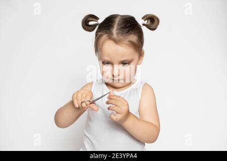 Primo piano ritratto di piccola cute concentrata 3 anni ragazza ragazzina, indossando top bianco e con stile ponytails capelli, tagliando il suo dito Foto Stock