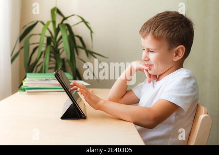 Carino scolaro che fa i compiti con il tablet alla scrivania. Bambino che utilizza gadget per lo studio e il gioco. Homeschooling durante la quarantena Foto Stock