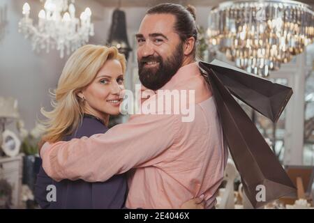 Felice bel bell'uomo maturo sopportato abbracciando la sua splendida moglie, tenendo borse per lo shopping sorridente alla macchina fotografica. Bella coppia matura sorridente allegro un Foto Stock