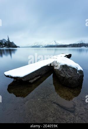 Vecchio molo di cemento coperto in uno strato di neve bianca su una tranquilla mattina invernale di Derwentwater nel Lake District, Regno Unito. Foto Stock