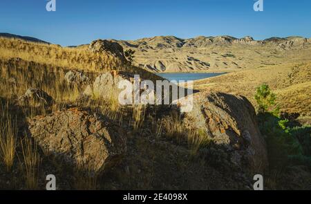 Paesaggio arido con massi, erbe e colline ai piedi delle Montagne Rocciose con scorcio di lago artificiale sotto il cielo blu vicino a Cody, Wyoming, Stati Uniti. Foto Stock