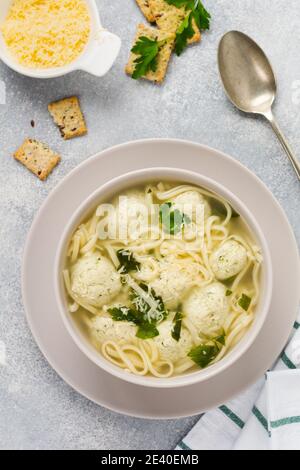 Ciotola di zuppa italiana tradizionale con polpette di pollo e pasta d'uovo, parmigiano, prezzemolo su sfondo grigio. Cucina italiana tradizionale. Foto Stock