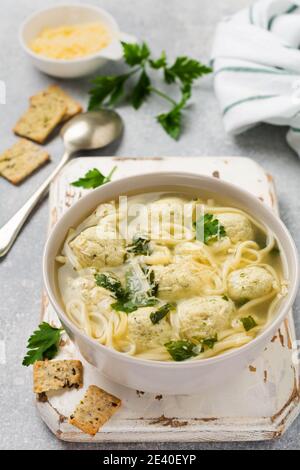 Ciotola di zuppa italiana tradizionale con polpette di pollo e pasta d'uovo, parmigiano, prezzemolo su sfondo grigio. Cucina italiana tradizionale. Foto Stock