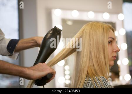 Profilo corto girato di una donna dai capelli biondi che ha i suoi capelli styled allo studio di bellezza. Professionale parrucchiere che utilizza l'asciugacapelli sul lavoro, styling capelli Foto Stock