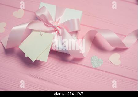 Scatola regalo bianca con arco in raso rosa e nastro su sfondo di legno rosa. Per il tuo testo è inclusa la tag mockup per matrimoni. Utilizzabile per San Valentino Foto Stock