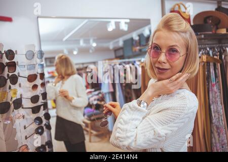 Primo piano di una donna elegante capelli biondi cercando occhiali rosa, sorridendo alla macchina fotografica, opy spazio. Allegra cliente femminile che si diverse a fare acquisti in Cloth Foto Stock