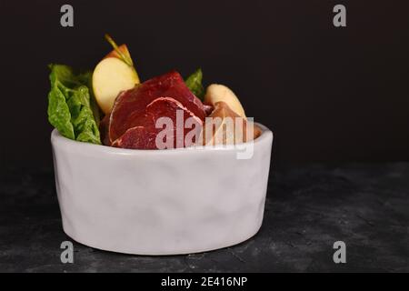 Ciotola del cane riempita con miscela di alimento crudo biologicamente appropriato contenente pezzi di carne, pesce, frutta e verdure su scuro Foto Stock